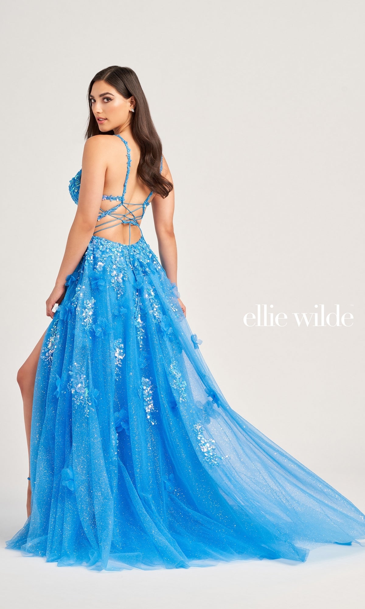 A woman models the Long Glitter Prom Ball Gown EW35047 by Ellie Wilde, featuring floral details, a lace-up back, striking blue glitter fabric, a high slit, and a long train against a plain light background.